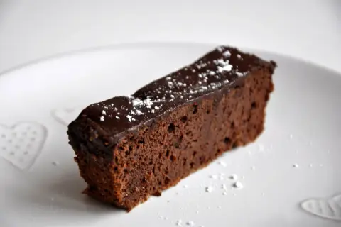Wilgotny brownie z fasoli, polany czekoladą i posypany cukrem pudrem, na białym talerzu z serduszkami.