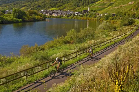 Zwei Radfahrer genießen die idyllischen radwanderwege deutschland entlang eines Flusses mit Weinbergen und einem Dorf im Hintergrund.