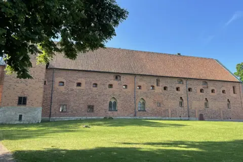 Imponerande tegelbyggnad med tegeltak, en del av Vadstena kloster, omgiven av grönskande gräsmatta och träd under en klarblå himmel.