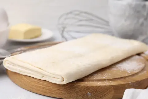 Gotowe ciasto francuskie na desce, obok masło i trzepaczka. Sprawdź, jaka jest ciasto francuskie cena!