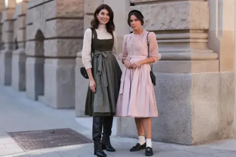 Zwei Frauen posieren vor einer Steinfassade. Eine trägt ein grünes Dirndl mit weißer Bluse und schwarze Stiefel.