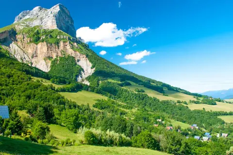 Vue panoramique d'une montagne imposante, le GR 965, dominant une vallée verdoyante parsemée de maisons. Le ciel bleu est parsemé de quelques nuages blancs.