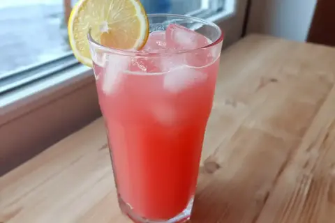 Ett glas rosa rabarberlemonad med is och en citronskiva, redo att avnjutas vid fönstret.