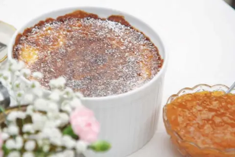 En gyllene, pudrad ostkaka i en vit form, bredvid en skål med aprikos sylt. Perfekt för ett ostkaka recept.