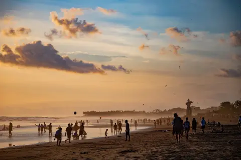 Zachód słońca na plaży Bali. Ludzie spacerują, grają w piłkę i puszczają latawce. W tle widać palmowy las.