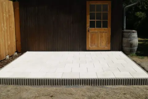 Nowy taras betonowy z jasnych płyt, otoczony ozdobnym krawężnikiem, przy drewnianej szopie z beczką na deszczówkę.