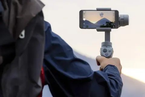 Osoba trzyma smartfon na gimbala, nagrywając piękny krajobraz z górami i zachodzącym słońcem.