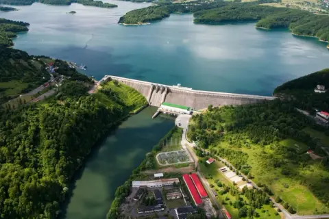Ogromna zapora na sanie spiętrza turkusową wodę jeziora otoczonego zielonymi wzgórzami. Widać budynki elektrowni i infrastrukturę.
