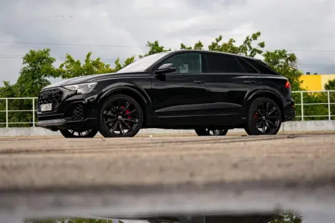 Czarny Audi Q9 z czerwonymi zaciskami hamulcowymi, odbijający się w kałuży. Sprawdź audi q9 cena!
