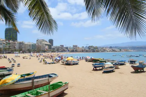 Solig strand i Las Palmas med palmer, båtar på sanden och en livlig stad i bakgrunden.