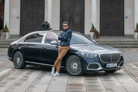 Elegancki mężczyzna opiera się o lśniącego Maybacha S580 przed monumentalnym budynkiem.