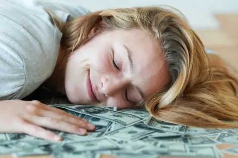 Uśmiechnięta kobieta śpi na stosie banknotów dolarowych. Marzy o tym, czy polskie gwiazdy rzeczywiście biorą udział w międzynarodowych kumulacjach.