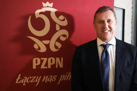 Uśmiechnięty prezes PZPN w garniturze stoi obok czerwonego banera z logo związku i hasłem "Łączy nas piłka".