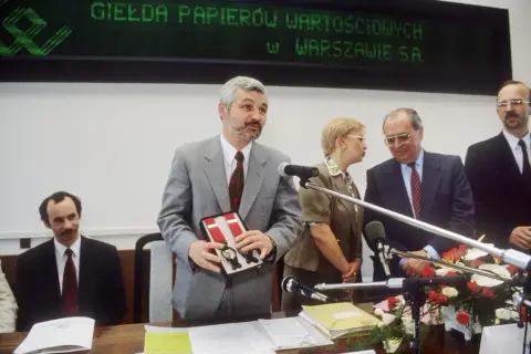 Uroczystość otwarcia, gdzie powstała pierwsza polska giełda. Mężczyzna w szarym garniturze prezentuje pudełko z szelkami.