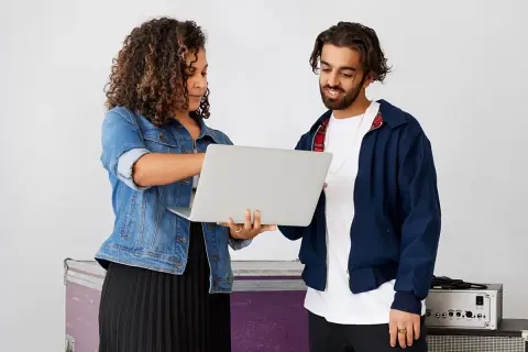 A woman and man review a laptop, discussing music artist management. They stand near stage equipment.