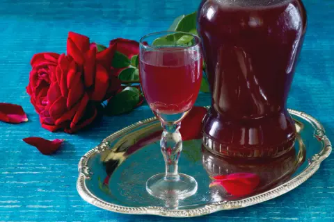 Aromatic nalewka z płatków róży w kieliszku i karafce, obok czerwona róża i płatki na srebrnej tacy.