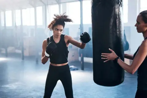 Woman training boxing for fitness at home, punching a heavy bag while a trainer holds it steady.