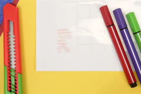 Un cutter rouge et trois feutres (rouge, violet, vert) sont prêts pour un projet créatif. Une **lettre noire** est visible sur le papier de fond.