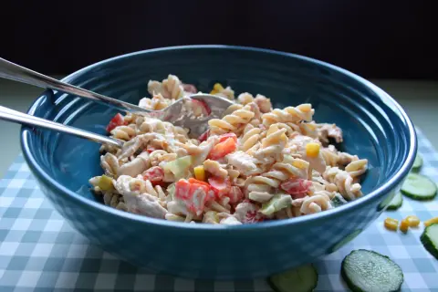 En stor blå skål fylld med krämig pastasallad med kyckling, tomat och majs. Två gafflar ligger ovanpå.