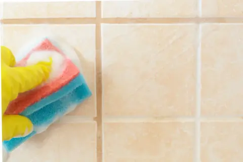 Mano con guanto giallo che pulisce le piastrelle del bagno con una spugna bagnata di schiuma.