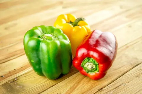 Tres pimientos de colores (rojo, verde y amarillo) sobre una mesa de madera. Los perros pueden comer pimiento, ¡una opción saludable!