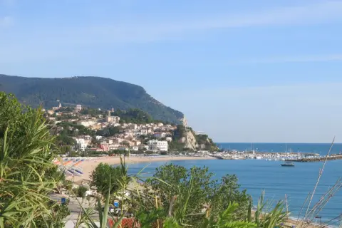 Vista panoramica della Riviera del Conero con un borgo arroccato su una collina, spiaggia attrezzata e un porto turistico.