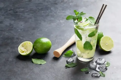 Orzeźwiający mohito smak: szklanka z lodem, limonkami i miętą, obok wyciśnięta limonka i tłuczek.