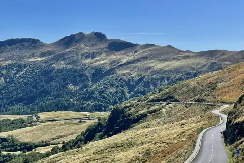 Route sinueuse dans les montagnes, avec des pâturages dorés et une forêt dense. Pas de Peyrol, un paysage grandiose sous un ciel bleu.