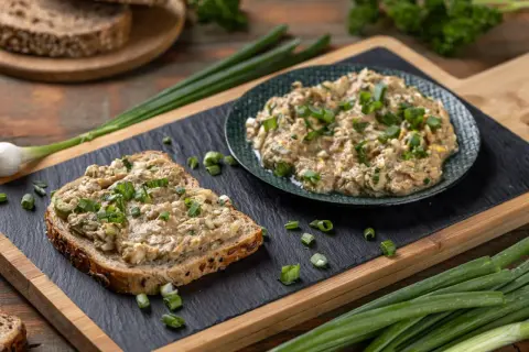 Pyszna pasta z tuńczyka na kromce ciemnego chleba, posypana szczypiorkiem. Obok talerz z pastą i świeże dymki.