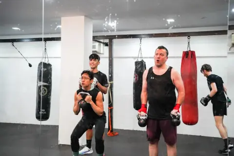 Men engage in boxing weight training, performing kettlebell squats and other exercises in a gym with punching bags.