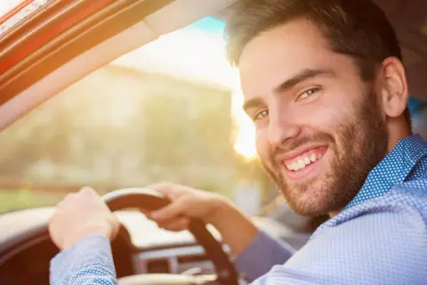 Un homme souriant au volant de sa voiture, prêt pour sa previsite contrôle technique. Le soleil brille, annonçant une journée sereine.