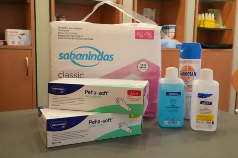 Pflegehilfsmittel Liste: Ein Paket Einlagen, zwei Schachteln Latex-Handschuhe, Desinfektionsmittel und ein Spray.