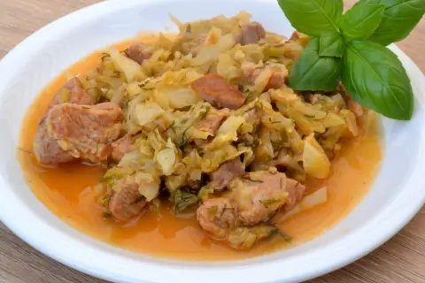 Pyszny bigos z młodej kapusty z kawałkami mięsa, podany na białym talerzu, ozdobiony gałązką bazylii.