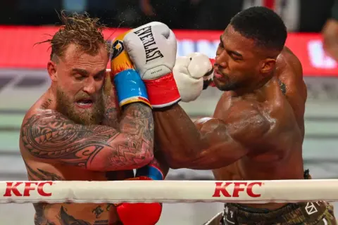 Two boxers grapple in the ring. One, with tattoos and wet hair, recoils from a punch. The other, muscular and focused, lands a blow. This is what heavyweight boxing looks like.