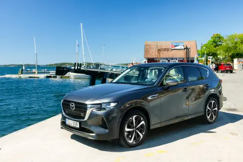 Mörkgrå Mazda CX-60 laddhybrid parkerad vid en brygga med båtar och en röd bil i bakgrunden. En solig dag för ett Mazda CX-60 laddhybrid test.
