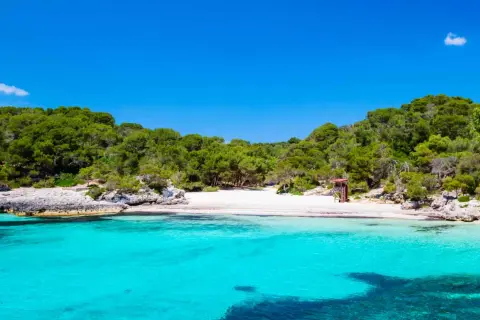 Aguas turquesas y arena blanca en la cala turqueta menorca, rodeada de pinos y rocas. Un paraíso balear.