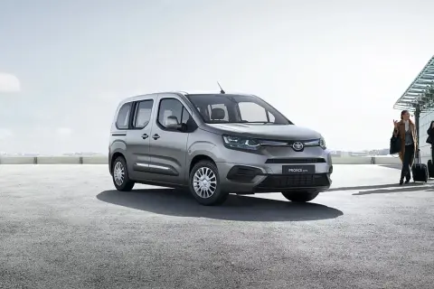 Srebrna toyota 7 osobowa czeka na pasażerów przy lotnisku. Ludzie z walizkami idą w stronę samochodu.