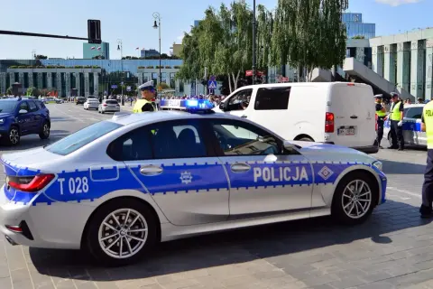 Radiowóz policyjny z włączonymi sygnałami świetlnymi na ulicy, obok policjanci.