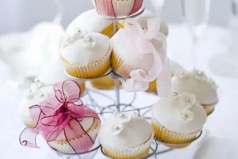 Zarte Muffins mit weißer Glasur, Blüten und Perlen, dekoriert mit rosa Bändern, perfekt für eine Hochzeit.