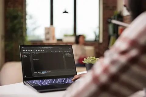 Programista pracuje nad kodem na laptopie, gdzie widać plik hosts. W tle siedzi osoba.