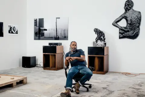 A man sits in an office chair in a gallery space, surrounded by art and audio equipment, a scene that feels like a snapshot of Arthur Jafa's creative process.