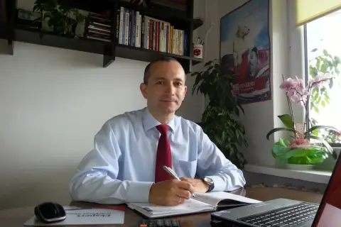 Mężczyzna w koszuli i krawacie siedzi przy biurku, pisząc w notatniku.