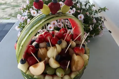 Koszyk z arbuza wypełniony owocami: truskawkami, winogronami, melonem i śliwkami. Idealne owoce na wesele, ozdobione malinami i borówkami.