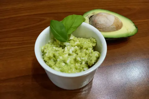 Pyszna pasta z awokado i jajka, udekorowana listkami bazylii, obok połówka awokado z pestką.