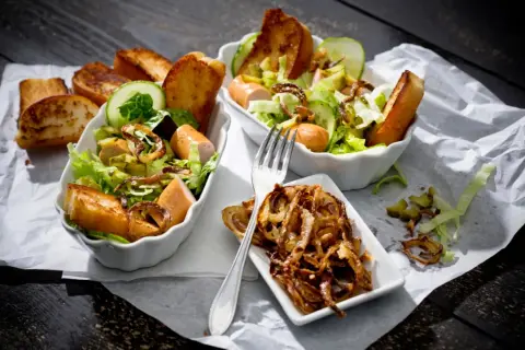 Zwei Schüsseln mit Hot Dog Salat, garniert mit Gurkenscheiben, geröstetem Brot und knusprigen Zwiebeln.