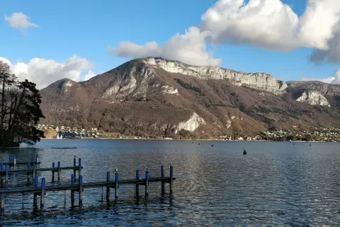 Vue de la webcam du lac d'Annecy, avec un ponton en bois et des montagnes majestueuses en arrière-plan.