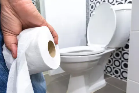 Dłoń trzymająca rolkę papieru toaletowego, w tle biała muszla klozetowa. Ktoś zmaga się z problemami z wypróżnianiem.
