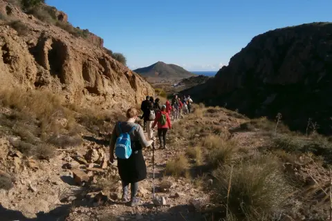 Senderismo en las **rutas de Cabo de Gata**. Un grupo de personas camina por un sendero rocoso y seco, con montañas a los lados y el mar al fondo.