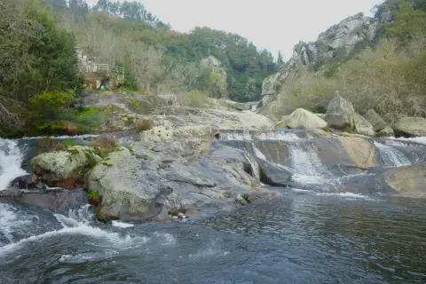 A fervenza de Segade, con auga caendo sobre rochas e un pequeno refuxio na ladeira verde.