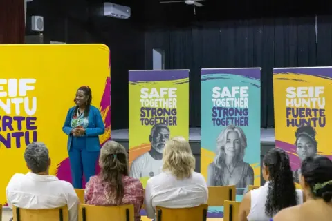Een spreker staat voor een gele banner met "SEIF HUNTU FUERTE HUNTU". Achter haar staan banners met "SAFE TOGETHER STRONG TOGETHER". Dit is een evenement voor hss sw jeugd.